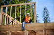 © anatoliy_gleb - Carpenter constructing wooden frame two-story house near the forest. Bearded man holding hammer, dressed in protective helmet and orange safety vest. Concept of ecological modern construction.