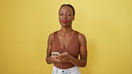 Wall Mural - Cheerful african american woman pointing and smiling while texting on her yellow smartphone against isolated background