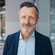 © contrastwerkstatt - Confident 55-Year-Old Businessman in Modern Shirt, Standing Before Sleek Glass and Concrete Wall, Gazing Relaxed into the Camera.