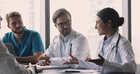 Wall Mural - Positive beautiful young Indian doctor woman talking to colleagues at meeting table, speaking and laughing, discussing career success, successful job case, medical cooperation