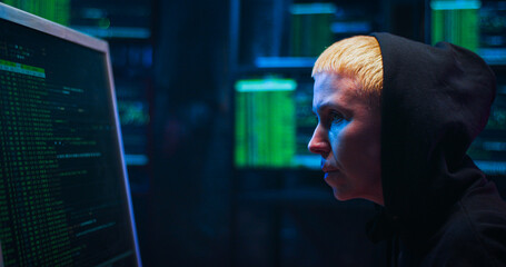 Side view on Caucasian woman cyber policewoman in dark hood sitting in room with monitors and typing on keyboard of computer. Female cyberpolice browsing online. Coding data. Save information concept