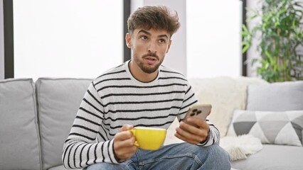 Sticker - Serious young arab man doubting over text message at home, sitting indoors, using his smartphone. relaxing with a drink of strong espresso coffee, lost in deep thought.