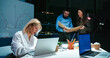 © ihorvsn - Caucasian young blonde woman talking on mobile phone and working at laptop computer in office at night shift. Male and female company co-workers cooperating on background. Lady speaking on cellphone.