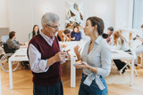 Fototapeta  - An animated conversation between an elderly gentleman and a younger woman, both standing, takes place in a bright workshop setting, surrounded by diverse attendees.