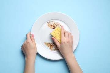 Sticker - Woman washing dirty plate with sponge on light blue background, top view