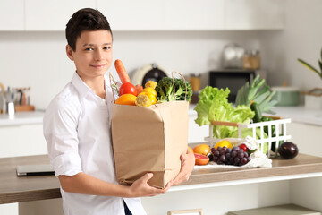 Sticker - Cute little boy holding paper bag with different vegetables in kitchen