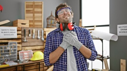 Poster - Cheerful young man, a professional carpenter, proudly sporting his beard, embodies good health - heart and hands on chest, showcasing a happy, confident aura in his carpentry workshop.