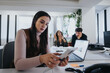 © qunica.com - A dedicated businesswoman calculates finance figures on a calculator in a bustling office environment with colleagues in the background.