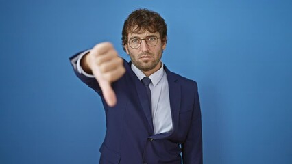 Canvas Print - Unhappy young man in business suit expresses anger with thumbs down gesture on isolated blue background. persistent sign of failure, sad portrait of blond adult?