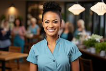 Happy Young Woman With Hair Up Wearing Blue Shirt With Smile Looking At Camera. Behind Her, There Are People Gathered Around Dining Tables With Chairs. There Are Potted Plants And Lights In Background
