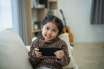 Wall Mural - Asian child girl looking using and touch mobile phone screen on couch sofa. Baby smiling funny time to use mobile phone. Too much screen time. Cute girl watching videos while tv, Internet addiction.