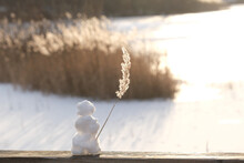 Little Snowman Standing On Bridge In Front Of A Frozen Lake Waving With A Reed In Backlight Of Sinking Winter Sun Like A Seaman On His Ship Waving Goodbye To Maybe The Last Snow.