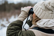 Woman dressed in winter clothes with gloves and knitted beanie looking for birds and animals through the binoculars in nature