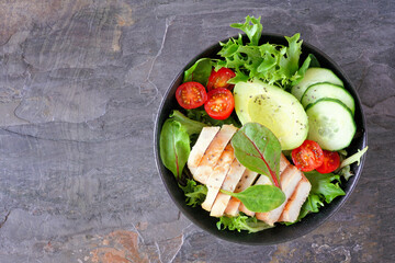 Wall Mural - Healthy homemade salad bowl with avocado and chicken. Above view on a dark slate stone background.