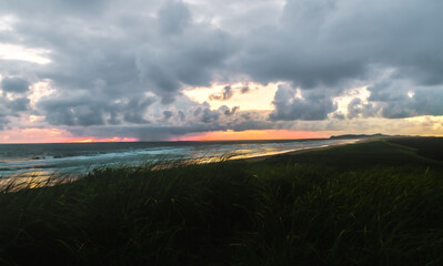 The sun sets behind rain clouds along the coast of the pacific ocean in Oregon casting light on the grassy beach area. 