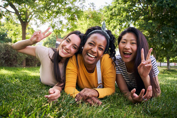 Wall Mural - Multi-ethnic group portrait smiling young women lying on grass in park. Three female laughing looking at camera making peace sign with hands enjoying weekend together. Generation z in sunny outdoors.