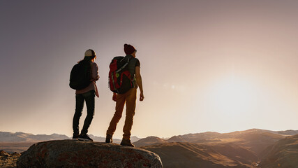 Wall Mural - two young active silhouettes of hikers with backpacks are standing on big rock and looks at sunset i