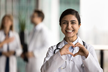 Wall Mural - Cheerful young Indian doctor woman making hand heart, looking at camera with happy toothy smile, showing romantic gesture of love, care, health protection, cardiology healthcare