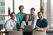 © fizkes - Diverse team of happy young doctors sitting and standing at laptop, looking at camera with toothy smiles, posing for group medic professional portrait. Medical practitioners promoting healthcare