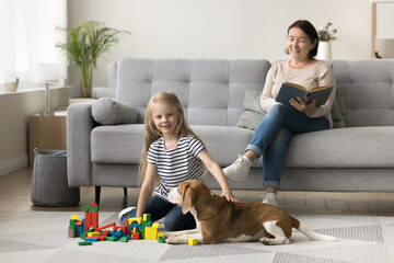 Wall Mural - Caring dog owner grandma watching little kid from couch, reading book. Granddaughter playing with construction blocks at home, sitting on floor, cuddling adorable pet, looking at camera, smiling