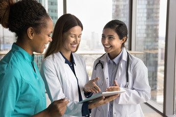 Wall Mural - Happy multiethnic young doctor women in white and blue uniforms using tablet for effective collaboration, pointing at screen, laughing, talking, discussing patient case, medical online service for job