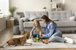 Positive caring mom and little kid playing with building blocks near dog on heating floor at home, constructing toy colorful tower from colorful, stacking cubes, enjoying family playtime with pet