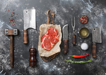 Wall Mural - Raw fillet of rib eye with kitchen meat utensils set on dark plate background. Cleaver,fork,knife with wooden tenderizer and various herbs and oil.