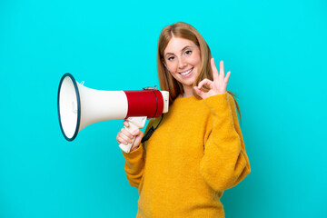 Wall Mural - Young redhead woman isolated on blue background holding a megaphone and showing ok sign with fingers