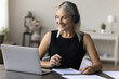 © fizkes - Happy pretty mature woman in headphones sit at desk, take notes, watch webinar on laptop, enjoy web course or online training, study remotely from home office, talking on video call to business client