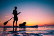 Silhouette of a man and a black dog on a Stand Up Paddle Board, SUP. Colorful sunset sky reflecting in the water. 