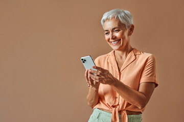 A beautiful stylish middle-aged woman stands on a beige background and holds a white smartphone with a smile. Advertising concert. Copy space.