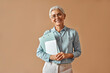 © HBS - Modern confident beautiful gray-haired mature business woman in blue blouse and glasses holding laptop and work folders while standing on beige background and smiling.