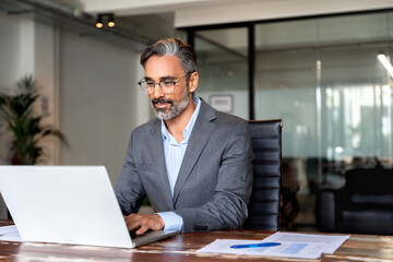Happy mature European business man ceo trader using computer, typing, working in modern office, doing online data market analysis, thinking planning tech strategy looking at laptop with copy space.