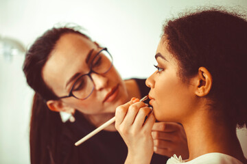 Young happy attractive african american woman visiting makeup artist in beauty salon, proffesional visagiste holding brush applying make-up on face of smiling black girl