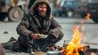 Homeless poor man sits and eats by the fire and warms himself in the cold