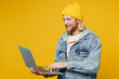 Side view young blond IT man wears denim shirt hoody beanie hat casual clothes hold use work on laptop pc computer typing message isolated on plain yellow background studio portrait Lifestyle concept