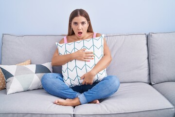 Poster - Young blonde woman hugging pillow sitting on the sofa in shock face, looking skeptical and sarcastic, surprised with open mouth