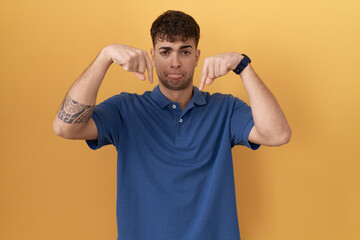 Canvas Print - Young hispanic man standing over yellow background pointing down looking sad and upset, indicating direction with fingers, unhappy and depressed.