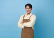 © NaMong Productions - Portrait young asian barista man wearing apron standing and smile arms crossed isolated on blue background, waitress or entrepreneur cheerful, small business or startup,