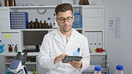 Wall Mural - Handsome young hispanic man, confident smiling scientist, comfortably working with touchpad at his high-tech lab, advancing in the world of science and medicine
