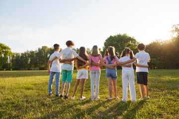 Group of teenage children look at sunset together during summer walk in green park. Rear view of kids standing in row hugging each other and looking in one direction. Concept of children's friendship.