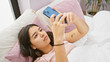 © Krakenimages.com - Young hispanic woman relaxing in bed with smartphone, sunlight illuminating cozy bedroom space.
