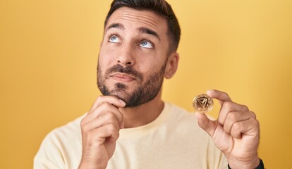 Wall Mural - Handsome hispanic man holding tron cryptocurrency coin serious face thinking about question with hand on chin, thoughtful about confusing idea