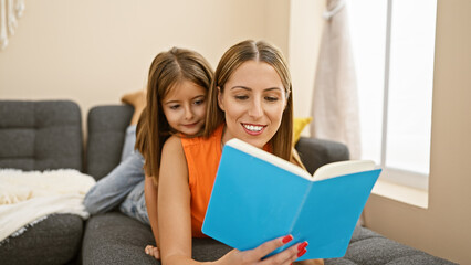 A smiling woman and her daughter enjoying reading a blue book together on a cozy sofa in their living room.