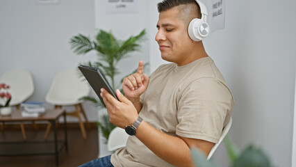 Canvas Print - Chill young latin man enjoying catchy songs with headphones, sitting comfortably in a waiting room, confidently smiling at camera on his touchpad