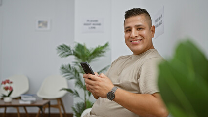 Poster - Cheerful young latin man happily texting away on his smartphone while sitting comfortably in a waiting room chair