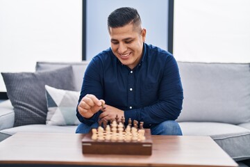 Wall Mural - Young latin man playing chess sitting on sofa at home