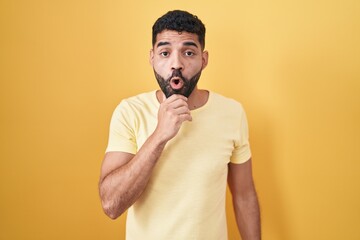 Sticker - Hispanic man with beard standing over yellow background looking fascinated with disbelief, surprise and amazed expression with hands on chin