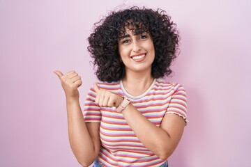 Sticker - Young middle east woman standing over pink background pointing to the back behind with hand and thumbs up, smiling confident