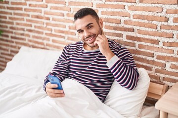 Poster - Young hispanic man using smartphone sitting on bed at bedroom